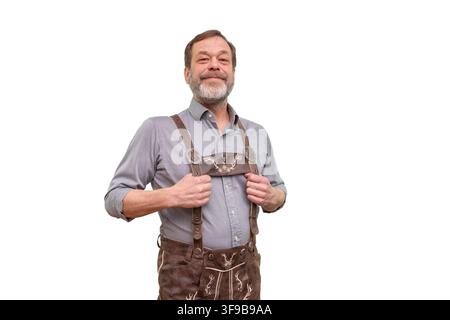 A smiling mature man stands in traditional lederhosen and a gray shirt while holding the suspenders against a clean light background. Stock Photo