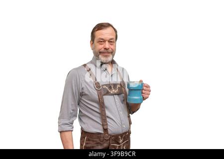 A mature bearded man wearing lederhosen stands facing the camera while holding a blue mug against a plain background. Stock Photo