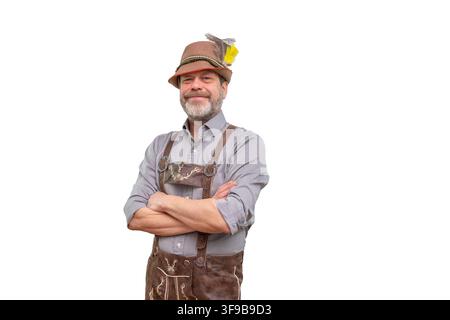 A smiling mature man wearing a Bavarian hat and traditional lederhosen stands with folded arms isolated on a white background. Stock Photo