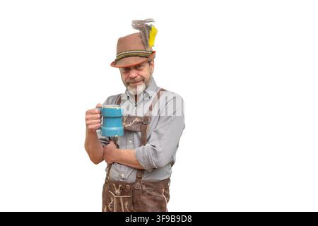 A bearded man in traditional Bavarian-style clothing stands isolated on a white background while holding a blue beer mug and looking at the camera. Stock Photo