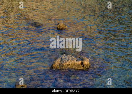 A serene natural scene featuring rocks partially submerged in flowing water, with the surface displaying gentle ripples and reflections, creating a tr Stock Photo