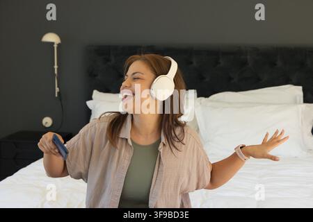 African American woman sitting on bed wearing white headphones, holding phone, smiling and singing Stock Photo