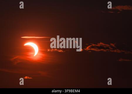 View of a partial solar eclipse with the sun partially obscured by the moon, set against a dark, dramatic sky with glowing orange clouds in Merikula, Stock Photo