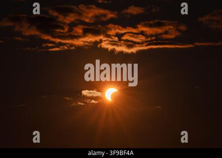 View of a partial solar eclipse with the sun partially obscured by the moon, surrounded by dark clouds and radiating sun rays in the sky in Merikula, Stock Photo