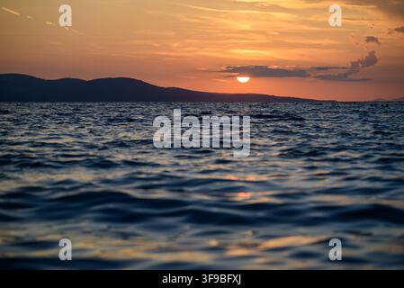 A tranquil sunset scene featuring a warm, glowing sun partially hidden behind a low-hanging cloud, casting golden light across the sky and reflecting Stock Photo