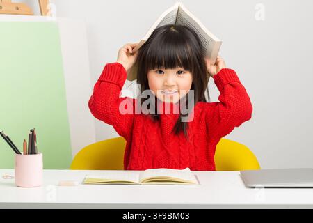 Playful Asian girl putting an open book on her head while sitting at desk. Stock Photo