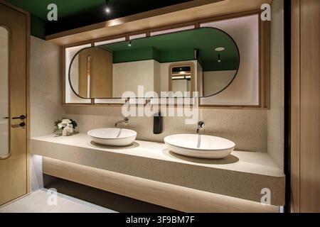 A sleek and contemporary public restroom interior featuring a beige terrazzo vanity counter with dual white round vessel sinks. A large modern oval Stock Photo