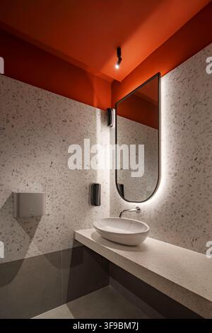 A modern commercial restroom design featuring a bold orange ceiling contrasting with grey terrazzo walls. The space includes a floating terrazzo. Stock Photo