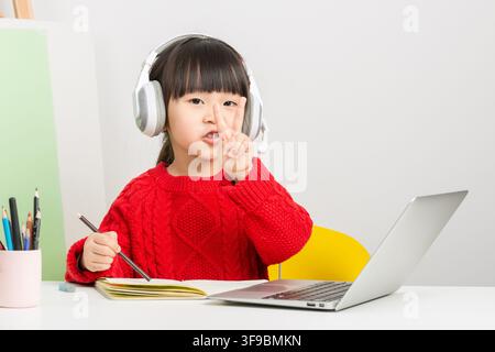 Smart Asian girl with headphones gesturing victory sign while studying with laptop online. Stock Photo