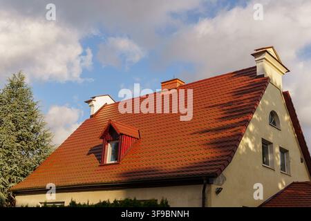 House featuring a red clay tile roof, a small dormer window, and a chimney, set against a blue sky with soft clouds, suburban residential architecture, classic roofing materials, real estate exterior Stock Photo