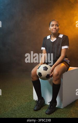 African female soccer player sitting on block on turf holding soccer ball under blue-orange lights Stock Photo