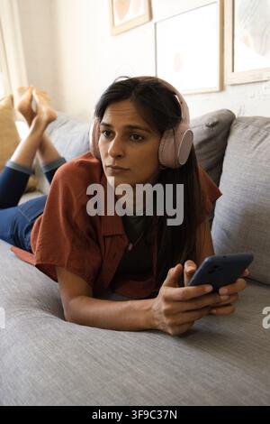 Adult Indian woman lying on sofa in living room, wearing pink headphones, holding blue smartphone Stock Photo