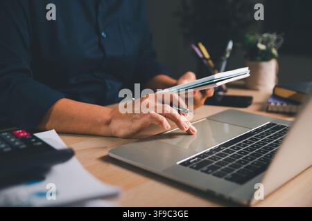 Business professional holding a notebook while using a laptop at a modern workspace, representing project planning, digital productivity, remote work, Stock Photo