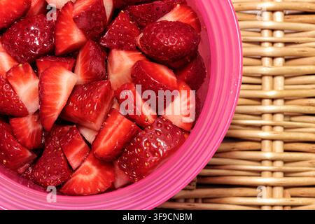 Top view close-up of fresh chopped strawberries served in a pink bowl on a wicker tray, healthy summer fruit concept Stock Photo
