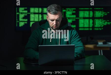 Hacker works on laptop at night. Hacker analyzes code screen in dark room. Hacker monitors network for cyber attack. Hacker targets data system with Stock Photo