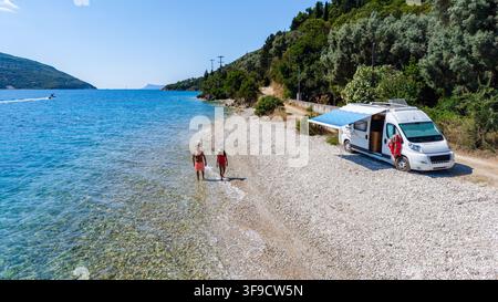 Crystal-clear waters embrace the serene shores of Kamari Beach in Lefkada, Greece. Couples stroll along the picturesque pebbled beach beside a lovely camper van. Stock Photo