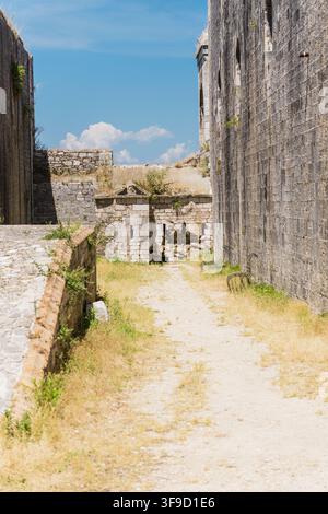 A sunlit dirt pathway passes between weathered stone walls and the ruins of an ancient fortress beneath a blue, partly cloudy sky Stock Photo