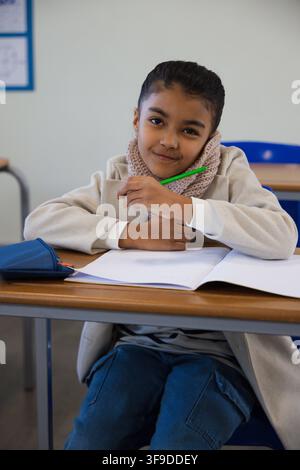Schoolgirl wearing pink scarf sitting at desk with open notebook in class, holding green pencil Stock Photo