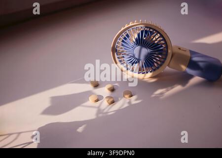 Handheld fan in blue and beige color next to round tablets on a light surface, with soft shadows created by natural light Stock Photo