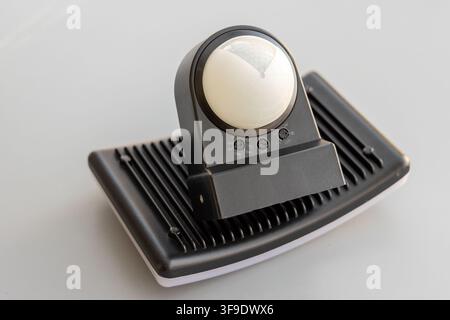 A black PIR motion sensor sits atop an LED floodlight on a white background Stock Photo