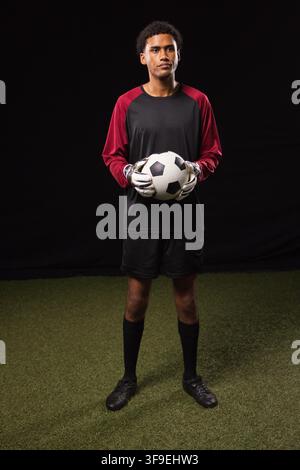 African American man standing on artificial turf in studio in keeper gloves and jersey holding ball Stock Photo