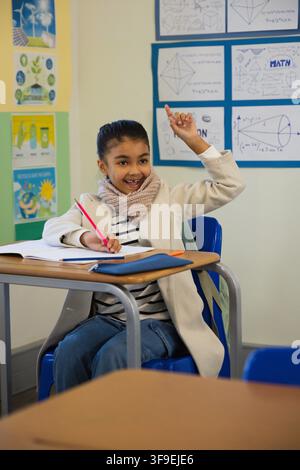 Female child sitting at school desk writing with red pencil over open notebook, wearing beige coat Stock Photo