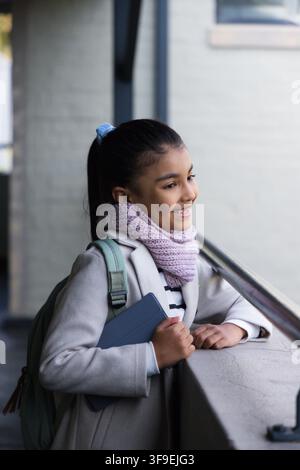 African American teen standing on balcony holding tablet wearing light-gray coat and lilac scarf. Backpack, ponytail, railing, concrete, device, stude Stock Photo
