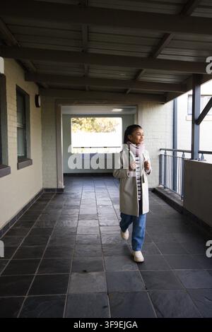 African teenage female walking along covered walkway on slate floor holding backpack, copy space Stock Photo