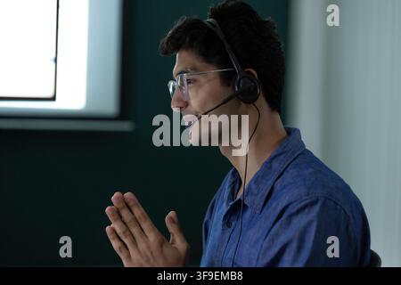 Indian male customer service agent wearing a headset while speaking with a client in an office, using a polite hand gesture during a call center Stock Photo