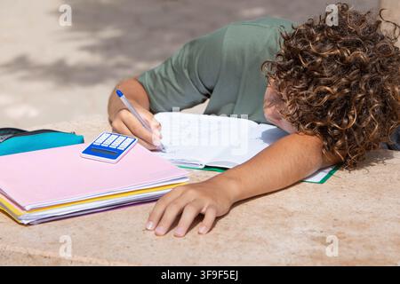 Tired Caucasian boy with curly hair falling asleep on his books while doing homework outdoors in a sunny park. Concept of student burnout Stock Photo