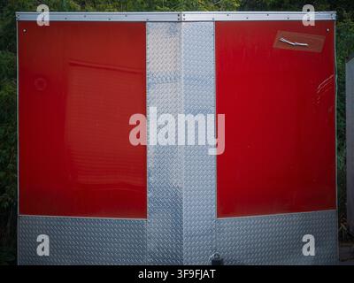 Red and silver diamond plate aluminum cargo trailer exterior wall panels outdoors with green trees Stock Photo