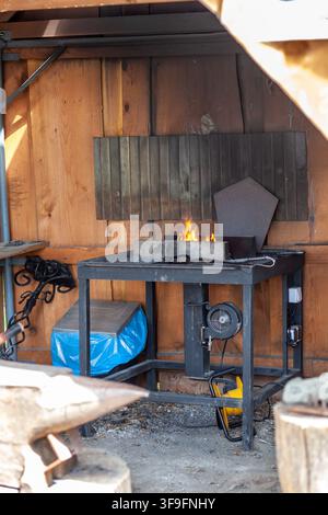 Blacksmithing workspace with forge, flames, and tools in a rustic wooden shed, featuring an anvil and metal sheets Stock Photo