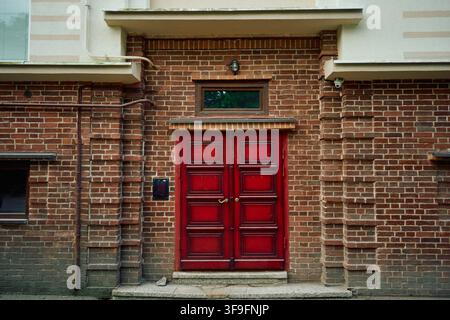 Deep red double wooden door recessed in aged brown brick facade with small transom window, exterior wall light, utility pipes and weathered urban Stock Photo