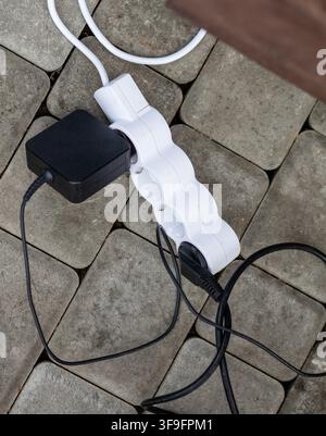 White power strip with multiple outlets and black charger on stone pavement, tangled cables visible in an outdoor setting Stock Photo