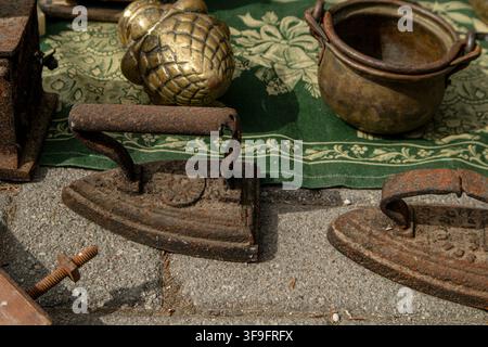 Heavy cast iron irons and brass vessels sit on a green textile surface at a busy flea market. People browse through various items on display. Stock Photo