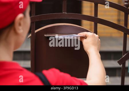 Mail carrier in red uniform delivering letters into a brown metal mailbox on a residential gate, close up view from behind Stock Photo