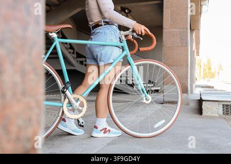 Young woman in denim shorts walking a turquoise fixed-gear bicycle with brown leather accents outdoors, Urban lifestyle and sustainable transportation Stock Photo