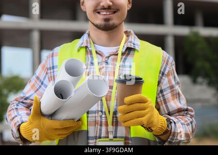 Smiling construction worker in safety vest holding blueprints and coffee cup at building site, the professional is on a building site, project develop Stock Photo