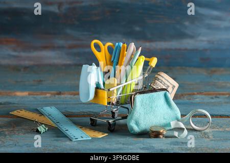 Miniature shopping cart filled with school supplies, money, and a coin purse on a rustic wooden surface Stock Photo