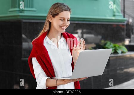 Smiling young woman waving during a video call on a laptop outdoors, remote work and communication concept, She is engaged in an online meeting or vir Stock Photo