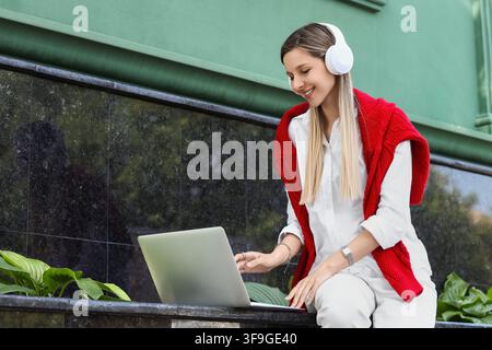 Smiling young woman with blonde hair wearing white headphones and a red sweater using a laptop outdoors, She is engaged in remote work or online learn Stock Photo