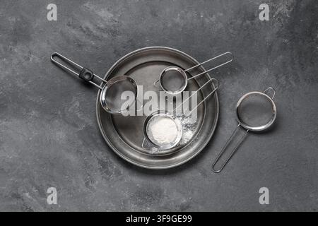 Stainless steel flour sifters and a metal tray with White powder on a dark gray concrete background Stock Photo