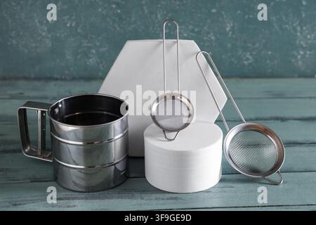 Stainless steel flour sifter and two small mesh strainers on white geometric pedestals with a blue wooden background, the kitchen tools are set agains Stock Photo