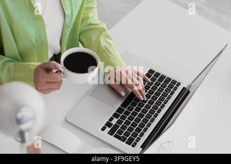 Womans hand typing on a laptop keyboard while holding a coffee cup, remote work and productivity Stock Photo