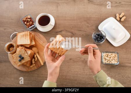 Young man spreading peanut butter on toast with spoon in kitchen, closeup Stock Photo