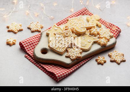 Delicious homemade christmas cookies shaped as snowflakes, gingerbread man, reindeer and houses lying on white cutting board on red checkered tablecloth with christmas lights Stock Photo