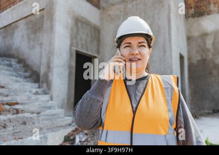 Woman in a construction site wearing a hard hat and orange safety vest. She is talking on a mobile phone with a serious expression Stock Photo