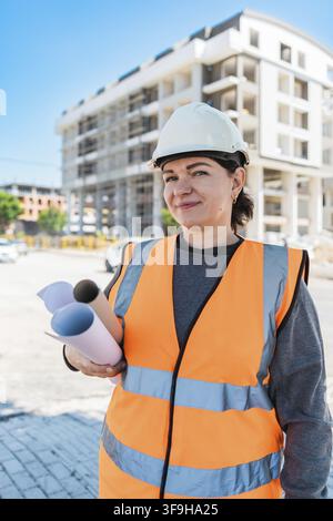 A young Hispanic woman wearing a hard hat and orange safety vest holds blueprints at a construction site with buildings in the background Stock Photo