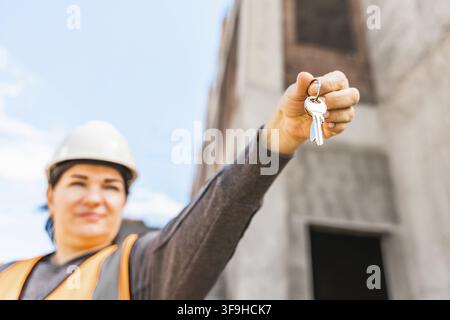 Close-up keys in hand. Woman in a hard hat and orange safety vest holds a set of keys in front of a construction site. The background shows unfinished Stock Photo