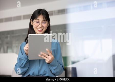 Young Asian professional woman reading e-mails, planning schedules, managing documents, working on digital tablet, preparing presentation, browsing bu Stock Photo